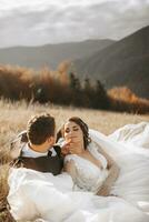 retrato do uma à moda noivo com uma noiva em uma fundo do outono seco grama. a conceito do uma rural Casamento dentro a montanhas, feliz boêmio recém-casados. a noiva e noivo estão deitado em a Relva foto