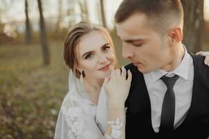 Casamento foto dentro natureza. a noivo senta em uma de madeira ficar em pé, a noiva carrinhos Próximo para ele, inclinado em dele ombro. Veja às a Câmera. retrato do a noiva e noivo