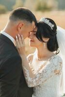 Casamento foto dentro natureza. a noivo dentro uma Preto terno e a noiva estão em pé dentro uma campo contra a fundo do montanhas, a noivo abraços dele Amado a partir de atrás e Beijos dela. retrato.