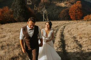 Casamento casal anda em dentro a montanhas. a noivo conduz a noiva de a mão. a vestir do a noiva desenvolve dentro a vento. foto