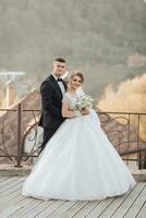 Casamento retrato. a noivo dentro uma Preto terno e a Loiras noiva estão de pé, abraçando, posando segurando uma ramalhete debaixo uma árvore. foto sessão dentro natureza. lindo cabelo e Maquiagem