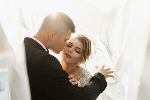 Casamento retrato. a noivo dentro uma Preto terno e a Loiras noiva estão abraçando, embrulhado dentro a da noiva véu. grandes vestir dentro a ar. foto sessão dentro natureza. lindo cabelo e Maquiagem