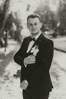 jovem elegante noivo dentro a floresta com uma flor na lapela dentro dele mão e uma arco gravata. Preto e branco foto