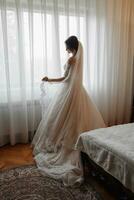 uma morena noiva dentro uma branco vestir e uma grandes véu poses em pé de a janela. lindo mãos. francês manicure. lindo cabelo e inventar. luz do dia foto