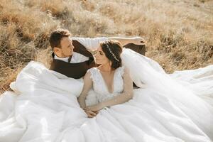 retrato do uma à moda noivo com uma noiva em uma fundo do outono seco grama. a conceito do uma rural Casamento dentro a montanhas, feliz boêmio recém-casados. a noiva e noivo estão deitado em a Relva foto