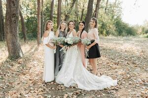 Casamento foto dentro natureza. uma noiva e dela damas de honra estão em pé dentro a bosques, sorridente, segurando seus buquês e olhando para dentro a Câmera lente. feliz Casamento conceito. emoções meninas