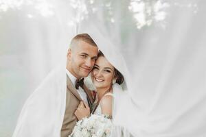 Casamento retrato do a noiva e noivo. feliz recém-casados suavemente abraço cada de outros debaixo a véu, pose, Veja às a Câmera e sorriso. à moda noivo. lindo jovem noiva foto