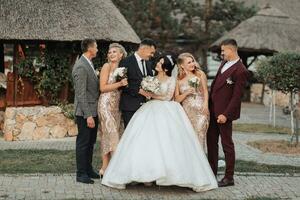 frente Visão do recém-casados em pé e abraçando, entre amigos. meninas dentro dourado vestidos aguarde buquês, a noiva dentro uma volumoso vestir, a noivo e dele amigos dentro se adequa. Casamento dentro natureza foto