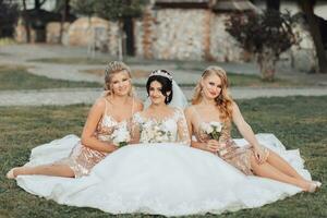 uma morena noiva dentro uma branco elegante vestir com uma coroa e dela Loiras amigos dentro ouro vestidos pose com buquês enquanto sentado em a grama. Casamento retrato dentro natureza, Casamento foto dentro uma luz tom.