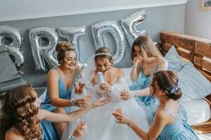 jovem damas de honra dentro azul seda vestidos beber champanhe dentro a da noiva sala. lindo mulheres a comemorar solteira festa sentado em cama e com champanhe. foto