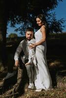 à moda modelo casal dentro a montanhas dentro verão. uma jovem menina dentro uma branco seda vestir carrinhos Próximo para dela marido contra a fundo do uma floresta e montanha picos. foto