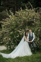 feliz jovem casal. Casamento retrato. a noiva ternamente abraçado a noivo, olhando às a Câmera contra a fundo do uma floração arbusto. Casamento ramalhete. Primavera Casamento foto