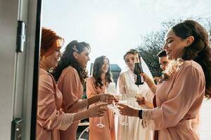 jovem damas de honra estão tendo Diversão e sorridente dentro Rosa seda vestes bebendo champanhe às a da noiva gazebo. lindo mulheres comemoro uma solteira festa, dentro uma gazebo e com champanhe. foto