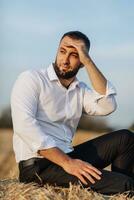 Casamento retrato foto. uma à moda noivo dentro uma branco camisa senta relaxado em uma feno fardo e parece para a lado, olhando gostar uma noiva. barbudo cara. estilo. foto