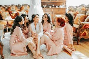 a damas de honra Veja às a sorridente noiva. a noiva e dela Diversão amigos estão a comemorar uma solteira festa em a chão perto a Casamento vestir dentro Coincidindo vestidos. noiva e amigos dentro a quarto foto