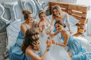 jovem damas de honra dentro azul seda vestidos beber champanhe dentro a da noiva sala. lindo mulheres a comemorar solteira festa sentado em cama e com champanhe. foto