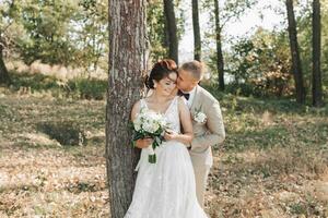 Casamento foto dentro natureza. a noiva e noivo estão em pé perto uma árvore, a noivo abraços dele Amado a partir de atrás e Beijos dela, ela sorrisos sinceramente. retrato. verão Casamento