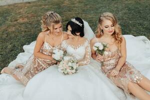 uma morena noiva dentro uma branco elegante vestir com uma coroa e dela Loiras amigos dentro ouro vestidos pose com buquês enquanto sentado em a grama. Casamento retrato dentro natureza, Casamento foto dentro uma luz tom.