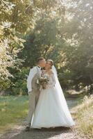 retrato do uma lindo casal dentro amor em seu Casamento dia. uma andar dentro a parque dentro a brilho do sol e vegetação. surpreendente Beijos e abraços do a noiva e noivo com uma ramalhete foto