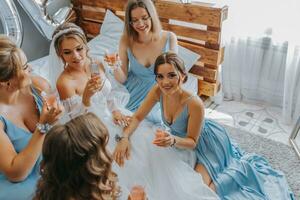 jovem damas de honra dentro azul seda vestidos beber champanhe dentro a da noiva sala. lindo mulheres a comemorar solteira festa sentado em cama e com champanhe. foto