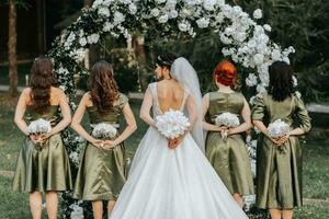 uma lindo noiva do detém uma Casamento ramalhete dentro dela mãos e parece às dela damas de honra quem estão segurando buquês com flores atrás seus costas foto