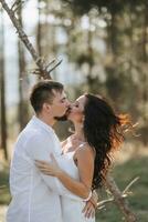 à moda noivo dentro branco camisa e fofa morena noiva dentro branco vestir dentro floresta perto Casamento de madeira arco. Casamento retrato do recém-casados. foto