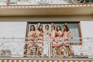 jovem damas de honra estão tendo Diversão e sorridente dentro Rosa seda vestes bebendo champanhe às a da noiva gazebo. lindo mulheres comemoro uma solteira festa, dentro uma gazebo e com champanhe. foto