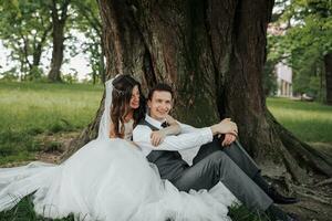 uma lindo noiva com grandes encaracolado cabelo dentro uma chique vestir abraços a noivo, sorrisos, olhando para dentro a lente debaixo uma grande árvore. retrato do a noiva e noivo. Primavera casamento. natural Maquiagem foto