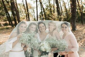 Casamento foto dentro natureza. a noiva e dela amigos estão em pé dentro a floresta segurando seus buquês, sorridente, coberto de a da noiva véu. Casamento retrato. emoções. lindo garotas.