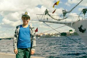 idade escolar Garoto vestindo do capitão boné posando de navio em água fundo foto