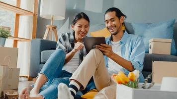 Feliz asiático jovem casal atraente homem e mulher usam tablet compras on-line móveis decorar a casa com pacote de papelão mover na casa nova. jovem casado asiático em movimento home shopper conceito on-line.