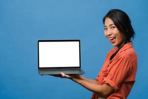 jovem asiática mostra a tela do laptop vazia com expressão positiva, sorri amplamente, vestida com roupas casuais, sentindo felicidade isolada sobre fundo azul. computador com tela branca na mão feminina. foto