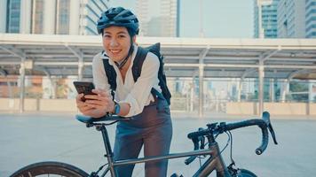 sorriso asiática empresária com mochila usar telefone inteligente olhar câmera na cidade ficar na rua com bicicleta ir trabalhar no escritório. garota esporte usa o telefone para o trabalho. comutar para trabalhar, viajante de negócios na cidade.