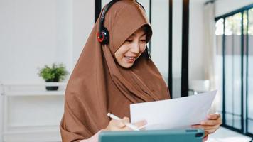 A senhora muçulmana asiática usa fone de ouvido usando tablet digital e fala com colegas sobre o relatório de venda em videoconferência enquanto trabalha em casa na cozinha. distanciamento social, quarentena para o vírus corona.