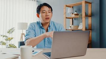 jovem empresário asiático usando laptop fala com colegas sobre o plano de videochamada enquanto inteligente trabalhando em casa na sala de estar. auto-isolamento, distanciamento social, quarentena para prevenção do vírus corona.