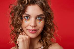 fechar-se foto do fofa jovem mulher, sorridente levemente, tocante suavemente dela bochecha, olhando para a Câmera, guardando mão dentro punho, posando em vermelho fundo