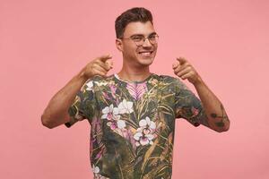 Boa olhando jovem homem sorridente amplamente e apontando dedos às Câmera, vestindo óculos e impresso camiseta, em pé sobre Rosa fundo, ser feliz e alegre foto