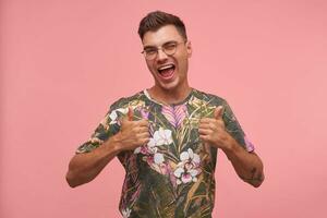 retrato do feliz jovem homem vestindo floresceu camiseta, manusear acima e piscando para Câmera, isolado sobre Rosa fundo, desfrutando vida foto