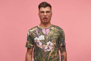 retrato do jovem cabelo curto homem olhando às Câmera, mostrando nojo, vestindo camiseta com floral impressão e copos, em pé sobre Rosa fundo foto