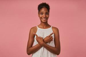 confuso jovem Castanho cabelos encaracolado Sombrio esfolado mulher fazendo careta confusamente dela face enquanto apontando com índice dedos dentro diferente instruções, isolado sobre Rosa fundo foto