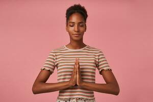 Boa olhando jovem lindo Castanho cabelos encaracolado fêmea com pão Penteado dobrando mãos juntos enquanto meditando com fechadas olhos, isolado sobre Rosa fundo foto