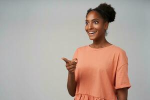 interior foto do jovem alegre bonita encaracolado fêmea com pão Penteado apontando surpreendentemente adiante com elevado indicador enquanto em pé contra cinzento fundo