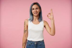 alegre jovem adorável de olhos azuis tatuado mulher com curto corte de cabelo mostrando bem feito placa enquanto olhando positivamente às Câmera, isolado sobre Rosa fundo foto