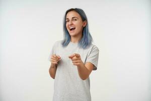 estúdio foto do jovem alegre tatuado senhora com curto azul cabelo guardando dela mãos elevado enquanto mostrando alegremente às Câmera com índice dedos, isolado sobre branco fundo