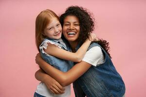 feliz atraente jovem senhoras com grandes cabelo sorridente Atenciosamente enquanto abraçando cada outro, desfrutando Tempo juntos enquanto posando sobre Rosa fundo dentro casual roupas foto