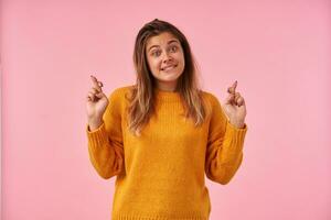 agitado jovem bonita Castanho cabelos senhora morder desconcertado dela lábio inferior e levantando mãos com cruzado dedos, vestindo casual roupas enquanto em pé sobre Rosa fundo foto