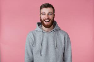 retrato do jovem feliz vermelho barbudo homem dentro cinzento capuz, parece feliz e amplamente sorrisos, carrinhos sobre Rosa fundo. foto