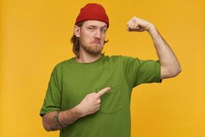 inseguro macho, bonito barbudo cara com loiro cabelo. vestindo verde camiseta e vermelho gorro. tem tatuagens. apontando com dedo e mostra dele bíceps. assistindo às a Câmera isolado sobre amarelo fundo foto