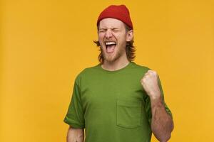 legal olhando macho, bonito barbudo cara com loiro cabelo. vestindo verde camiseta e vermelho gorro. apertar dele punho dentro excitação, celebração. ficar de pé com fechadas olhos, isolado sobre amarelo fundo foto