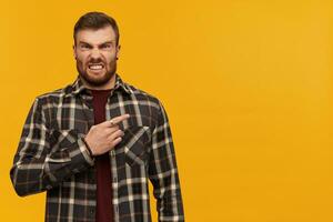 Bravo irritado jovem homem dentro xadrez camisa com barba em pé e apontando de dedo longe para a lado sobre amarelo fundo foto